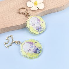 two keychains with anime characters on them sitting next to a flower and a piece of wood