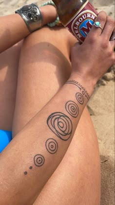 a woman sitting on the beach with a beer in her hand and tattoos on her arm