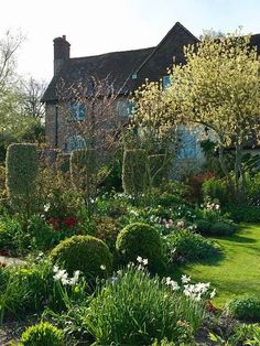 the garden is full of beautiful flowers and trees in front of a large house with blue shutters