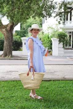 a woman in a blue dress and hat holding a straw bag on the grass near a tree