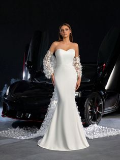 a woman standing in front of a black car wearing a white dress with feathers on it