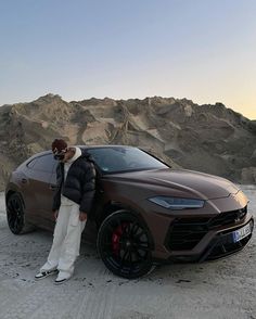 a man standing next to a brown sports car in the middle of desert with mountains behind him