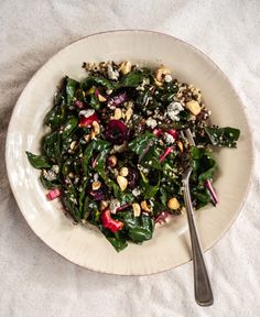 a white plate topped with spinach salad next to a fork and spoon on a table