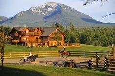 two people riding horses in front of a large log house with mountains in the background