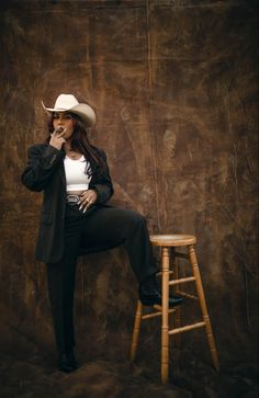 a woman sitting on top of a wooden stool wearing a cowboy hat and black pants