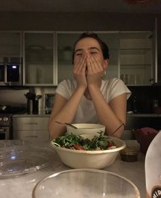 a woman covering her eyes while sitting at a table with a bowl of salad in front of her