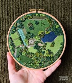 someone is holding up a small embroidery project in front of the camera, which shows an aerial view of a park