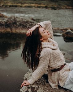 a woman sitting on top of a rock next to a river wearing a sweater and scarf