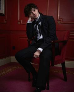 a man in a suit and tie sitting on a red chair with his hand to his face