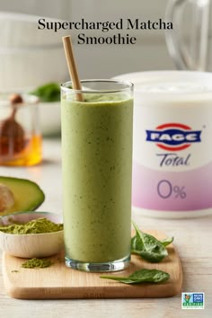 a green smoothie in a tall glass with a wooden spoon on the side next to an avocado and yogurt