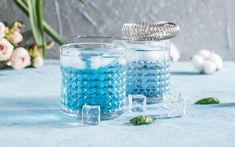 two glasses filled with blue liquid sitting on top of a table next to some flowers