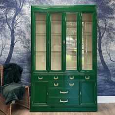 a green china cabinet sitting in front of a wall with trees painted on the walls