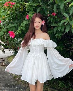 a woman in a white dress standing next to some bushes and pink flowers with her hands on her hips