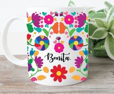 a white coffee mug with colorful flowers and the word bonita on it next to a potted plant