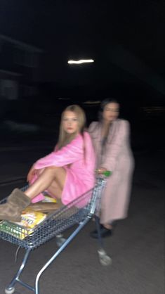 two women are sitting in a shopping cart at night, one is wearing a pink robe