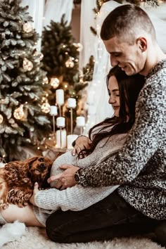 a man and woman sitting on the floor with their dog in front of a christmas tree