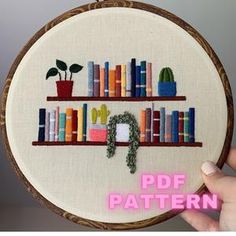 a hand holding up a cross stitch book shelf with books on it and a potted plant