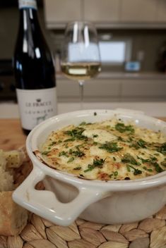 a white dish filled with food next to a bottle of wine and some slices of bread