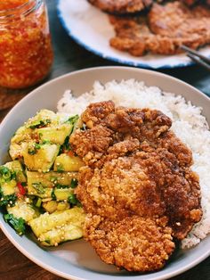 two plates with fried chicken, rice and vegetables on them next to a jar of sauce