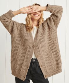 a woman wearing a cardigan sweater and jeans with her hands on her head, smiling