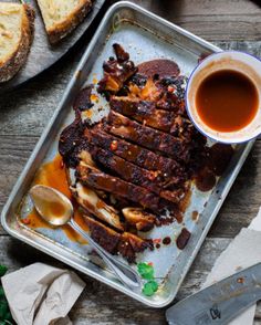 a pan with ribs and sauce on it next to bread, butter and spoons