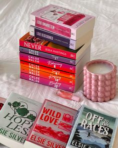 a stack of books sitting on top of a bed next to a pink cup and candle