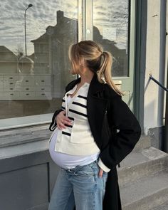 a pregnant woman standing in front of a window with her back to the camera, wearing jeans and a black coat