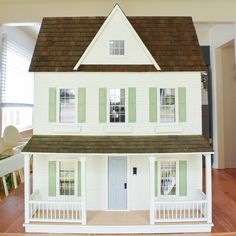 a doll house with green shutters on the front and side windows, sitting on a wooden floor