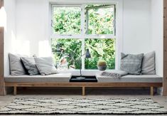 a window seat with pillows on it in front of a rug and table next to the window