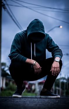 a man squatting on the ground with his hands in his pockets and wearing a hoodie