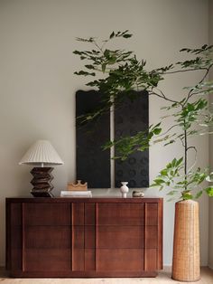 a wooden cabinet sitting next to a tall plant in a room with two paintings on the wall