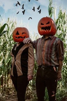 two people wearing pumpkin heads standing next to each other in front of cornfield with birds flying overhead