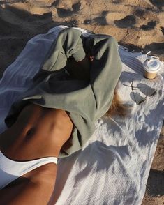 a woman laying on top of a white towel next to a cup and bottle in the sand