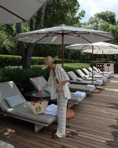 a woman in a white dress and straw hat standing next to lounge chairs with an umbrella