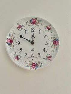 a white wall clock with flowers on it's face and numbers in the middle