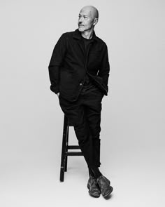 a man sitting on top of a stool in front of a white wall with his legs crossed