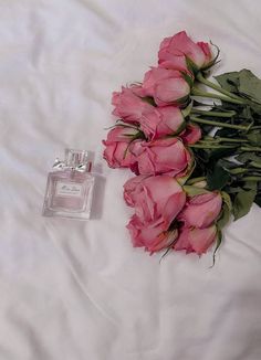 a bouquet of pink roses next to a bottle of perfume on a white bed sheet