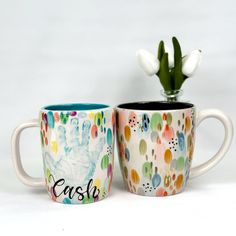 two coffee mugs with designs on them, one has a hand print and the other has a flower