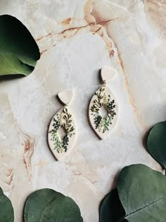 two white ceramic earrings with floral designs on them next to green leaves and marble background