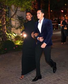 a man and woman dressed in black are walking down the street at night with their arms around each other