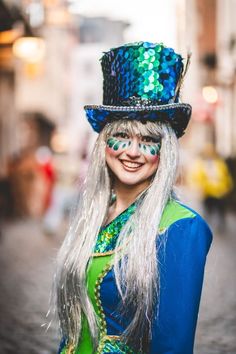 a woman with long white hair wearing a blue top hat and green dress is smiling at the camera