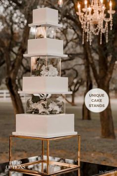 a white wedding cake sitting on top of a metal stand with chandelier hanging from the ceiling