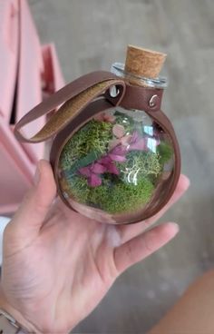 a hand holding a glass bottle filled with moss and pink flowers on top of a table