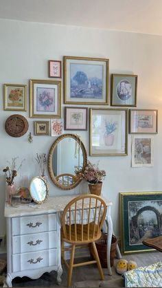 a white desk topped with a wooden chair next to a wall covered in framed pictures