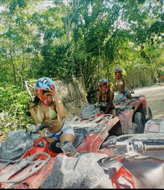 three people on four wheelers in the woods with trees and bushes behind them, one woman is talking on her cell phone