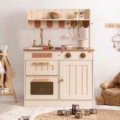 a wooden toy kitchen with lots of toys on the floor in front of it and a teddy bear sitting next to it