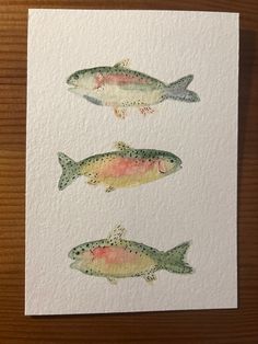 three watercolor paintings of fish on white paper with brown wooden table in the background