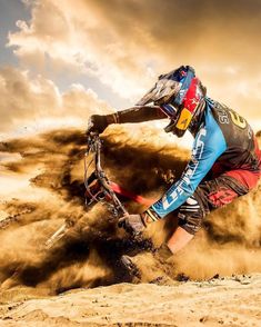 a man riding a dirt bike on top of a sandy ground covered in dust with clouds behind him