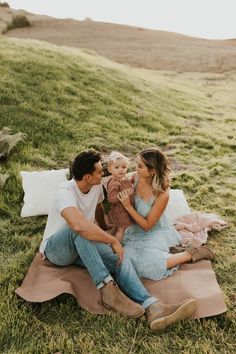 a man, woman and child sitting on a blanket in the grass with one holding a baby