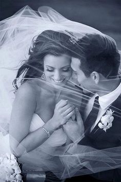 a bride and groom pose for a wedding photo with veil blowing in the wind over their heads